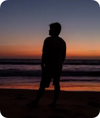 A silhouette of a person standing on a beach during sunset, with vibrant orange and purple hues in the sky reflecting on the ocean waves.